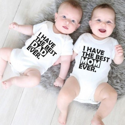 Two newborn babies lying on a furry surface, wearing white rompers with text 'I HAVE THE BEST MOM EVER' and 'I HAVE THE BEST DAD EVER' printed on them.