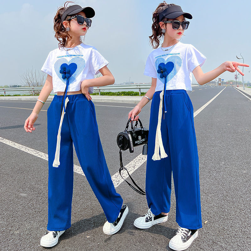 Two girls wearing matching outfits consisting of white tops with a heart graphic and blue wide-leg pants, one holding a bag and posing on a road surface.