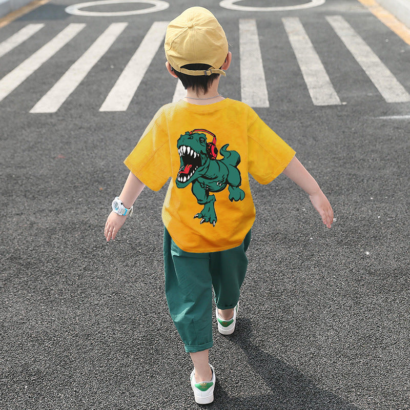 A child wearing a white short-sleeve shirt with a cartoon dinosaur print and green pants, walking away from the camera.