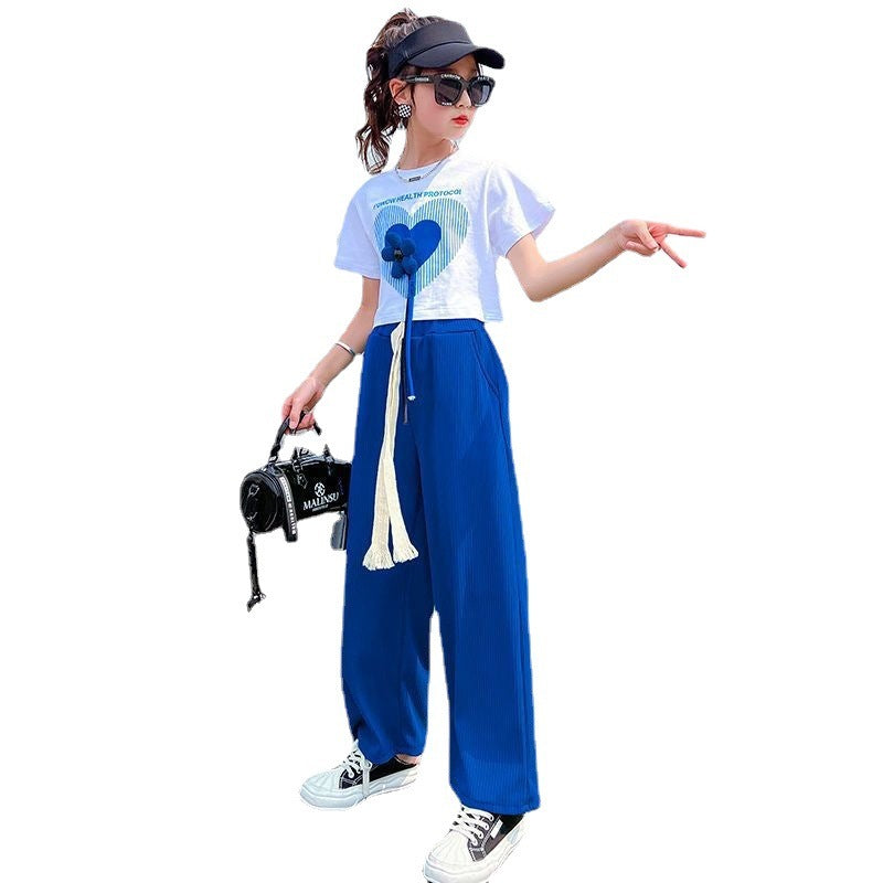 Two girls wearing matching outfits consisting of white tops with a heart graphic and blue wide-leg pants, one holding a bag and posing on a road surface.