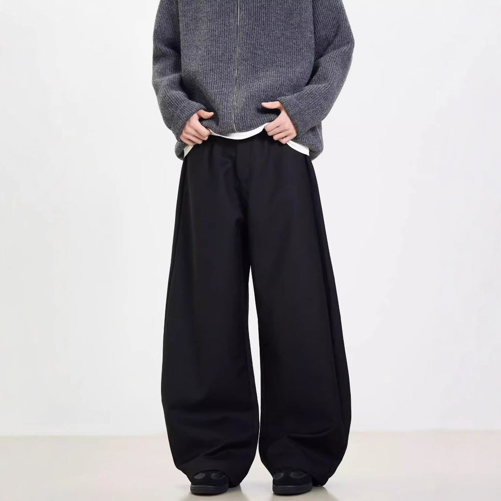 A person standing against a white background, wearing black wide-leg suit pants and a gray sweatshirt.