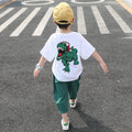 A child wearing a white short-sleeve shirt with a cartoon dinosaur print and green pants, walking away from the camera.