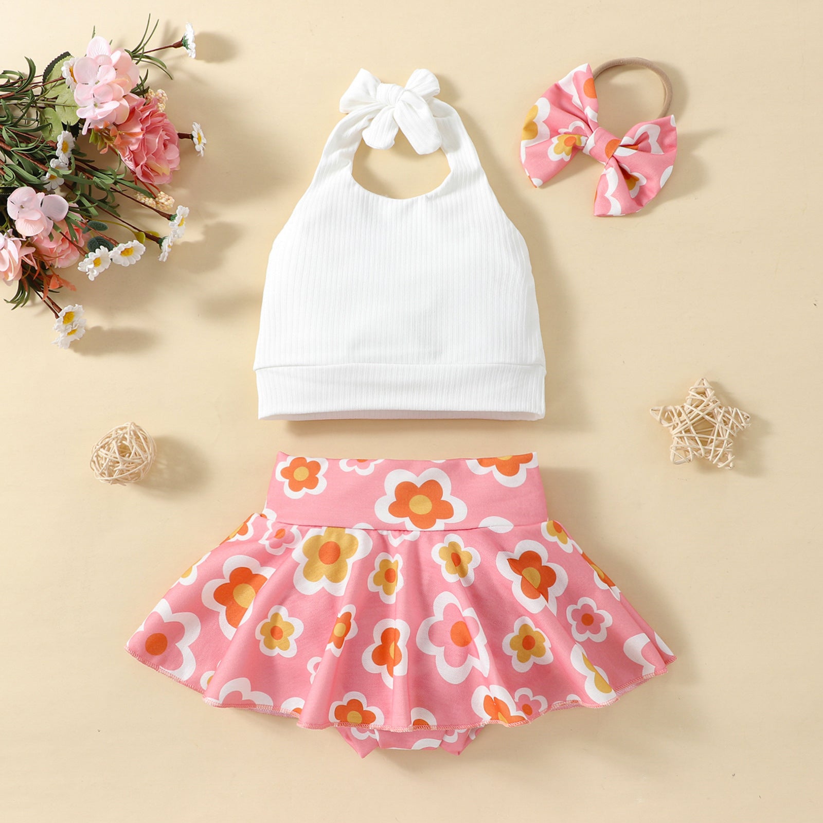 A set of children's clothing including a white sleeveless top with a bow at the neck, a pink skort with a floral pattern, and a matching hairband, displayed against a beige background with decorative elements like flowers and stars.
