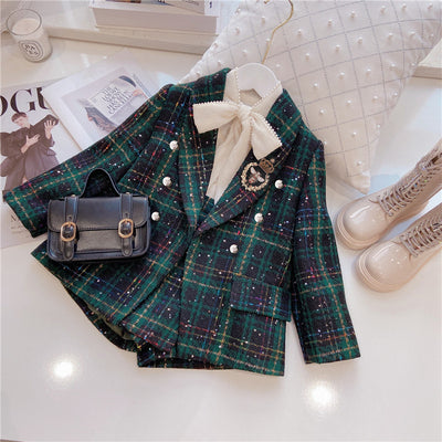 A children's two-piece suit with a green plaid pattern, consisting of a blouse with a bow tie and pants, accompanied by a small handbag and shoes.
