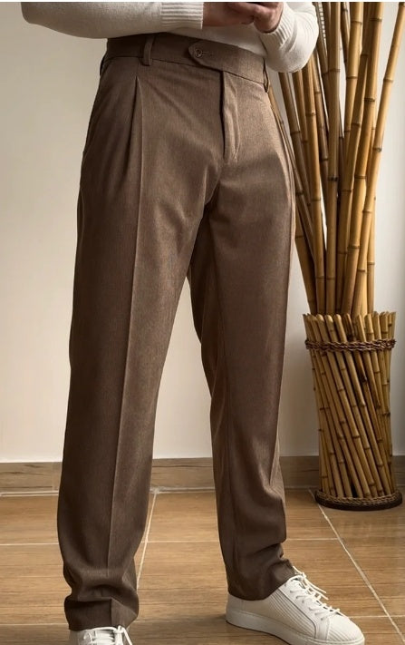 A person standing in a room with bamboo decor, wearing brown high-waisted slim fit trousers and white shoes.