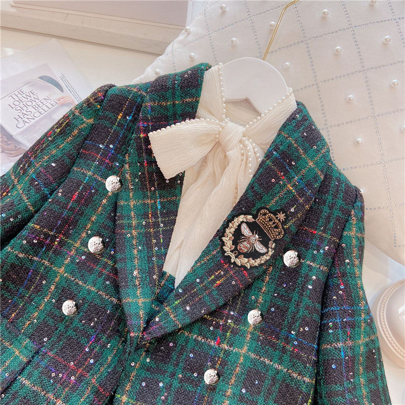 A children's two-piece suit with a green plaid pattern, consisting of a blouse with a bow tie and pants, accompanied by a small handbag and shoes.